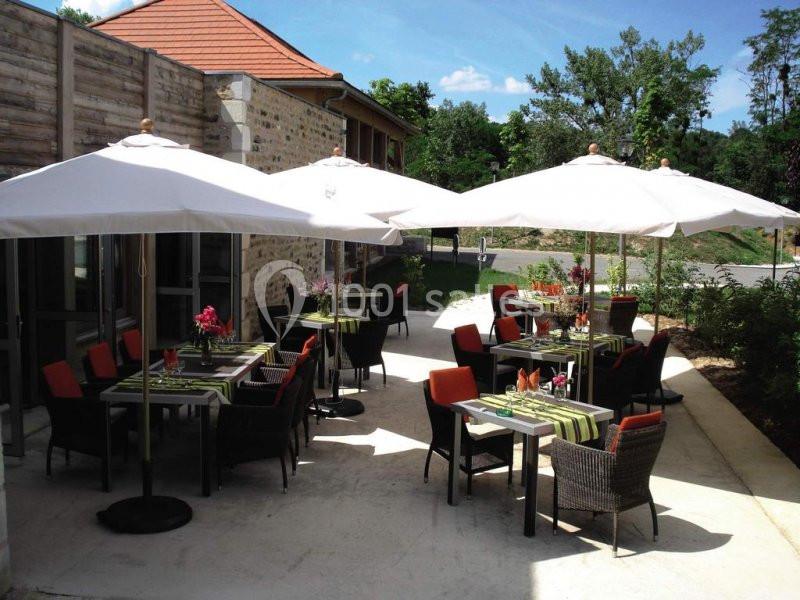 Terrasse extérieure avec tables dressées, chaises en osier et parasols blancs, entourée de verdure et bâtiments en pierre.
