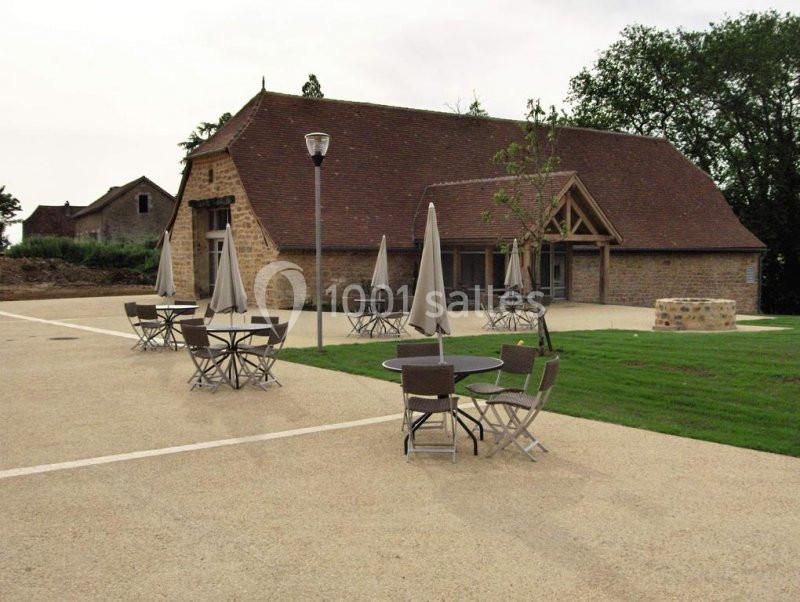Bâtiment en pierre avec toit en tuiles, entouré de tables et parasols sur une terrasse pavée, avec pelouse à proximité.