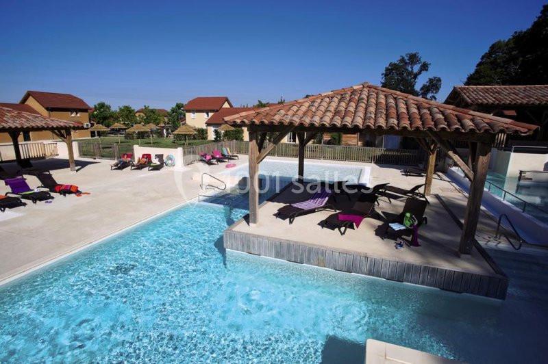 Piscine extérieure avec pergolas en bois, transats colorés et maisons en arrière-plan sous un ciel dégagé.
