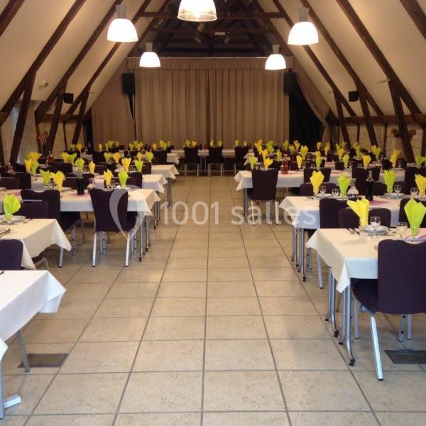 Salle de réception avec tables dressées, nappes blanches, serviettes jaunes pliées et chaises violettes.