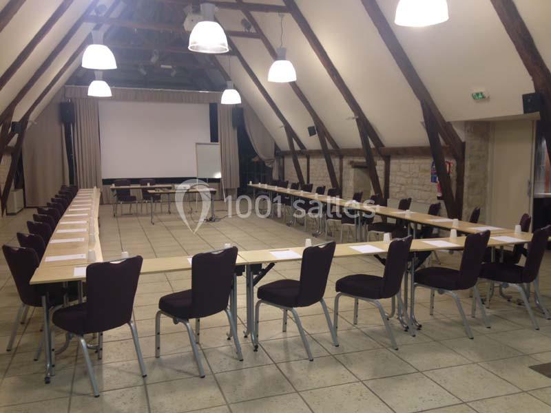 Salle de réunion spacieuse avec des tables disposées en U, des chaises alignées et un écran de projection au fond.