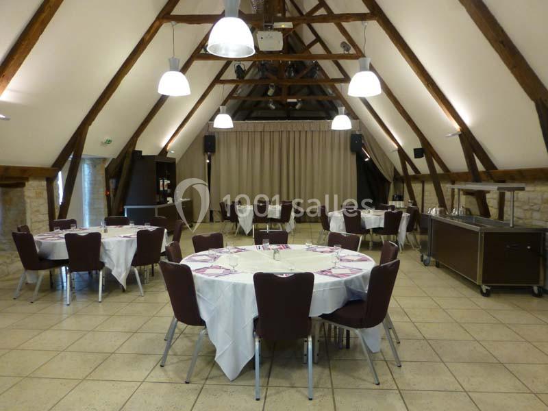 Salle de réception avec des tables rondes dressées, poutres apparentes et éclairage suspendu.