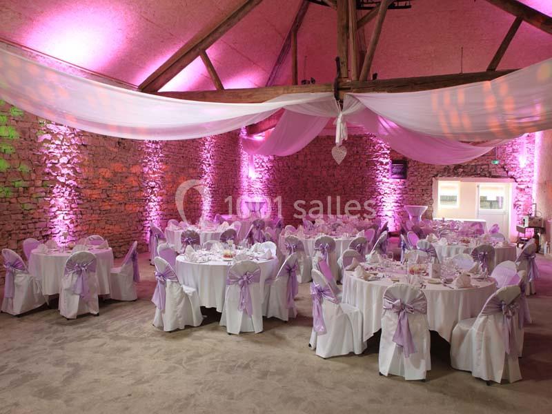 Salle de réception décorée avec des tables rondes, nappes blanches et nœuds roses, éclairée par des lumières tamisées.