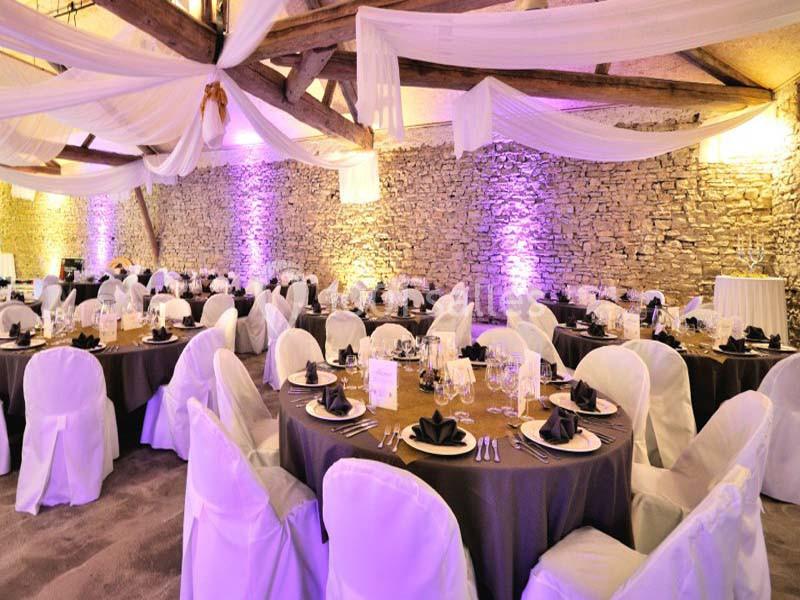 Salle de réception décorée avec des tables rondes dressées, nappes sombres et chaises blanches, éclairage violet.