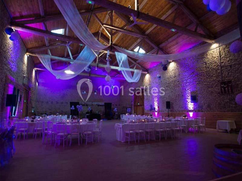 Salle de réception en pierre avec tables dressées, éclairage violet et décorations suspendues en tissu blanc.