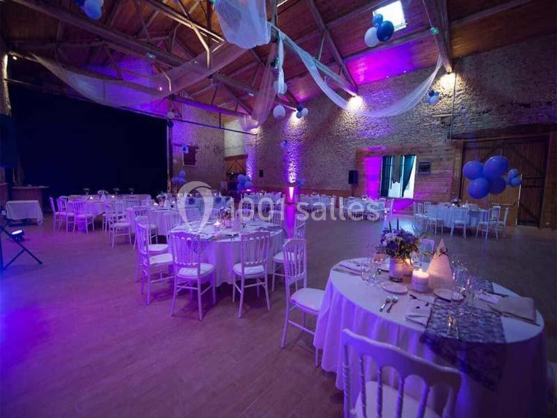 Salle de réception décorée avec des tables rondes, chaises blanches, éclairage violet et ballons suspendus.