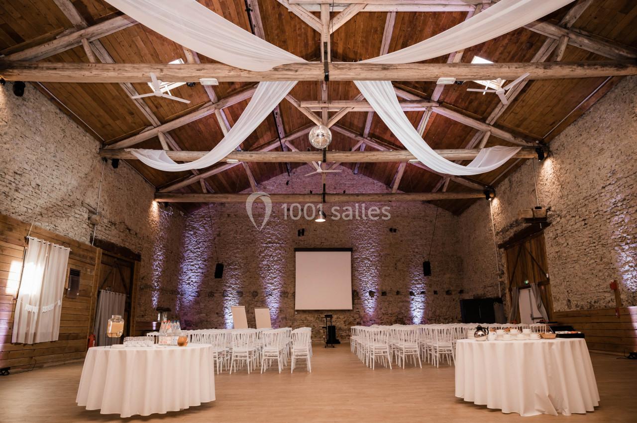 Salle en pierre avec plafond en poutres apparentes, décorée de voilages blancs, tables rondes et chaises blanches.
