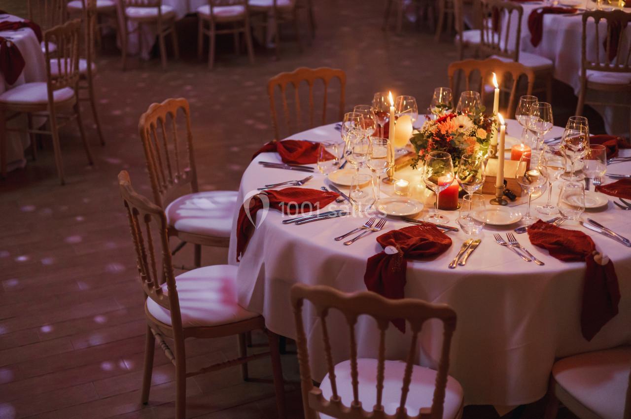 Table ronde élégamment dressée avec nappes blanches, serviettes rouges, bougies et centre de table floral.