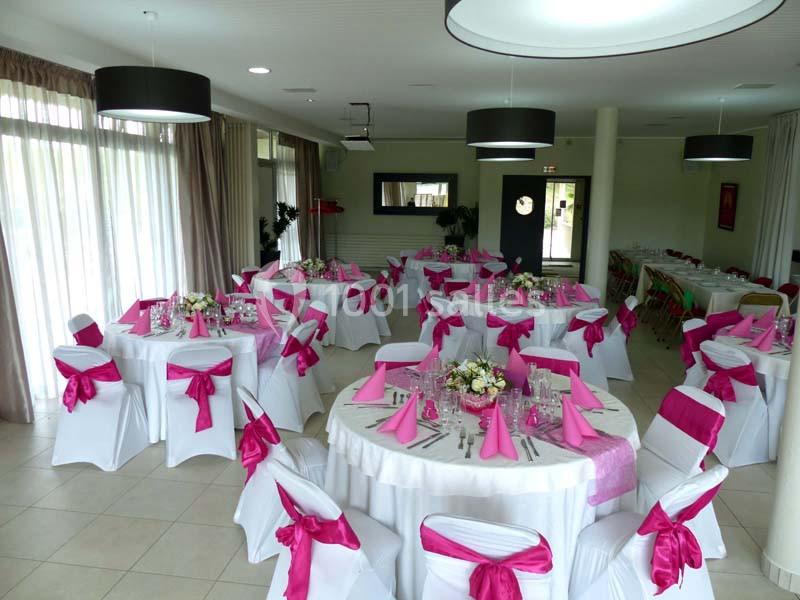 Salle de réception décorée avec des tables rondes, nappes blanches et nœuds roses sur les chaises.