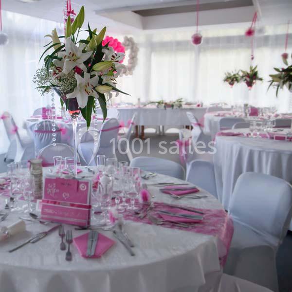 Salle de réception décorée en blanc et rose avec des tables dressées et des centres de table fleuris.