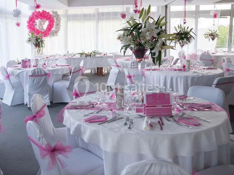 Salle décorée pour un événement avec des tables rondes dressées, ornées de nappes blanches et de décorations roses.