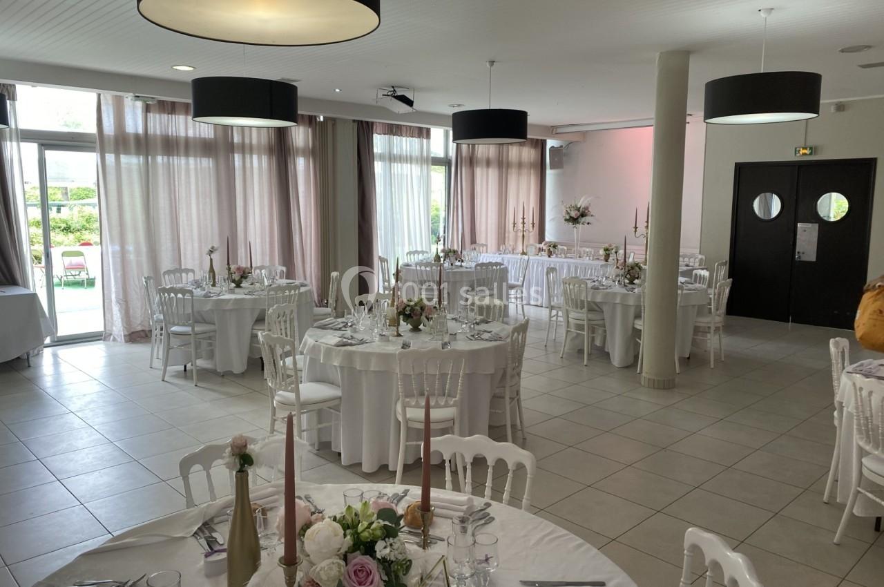 Salle de réception lumineuse avec tables rondes dressées, nappes blanches, décorations florales et grandes baies vitrées.