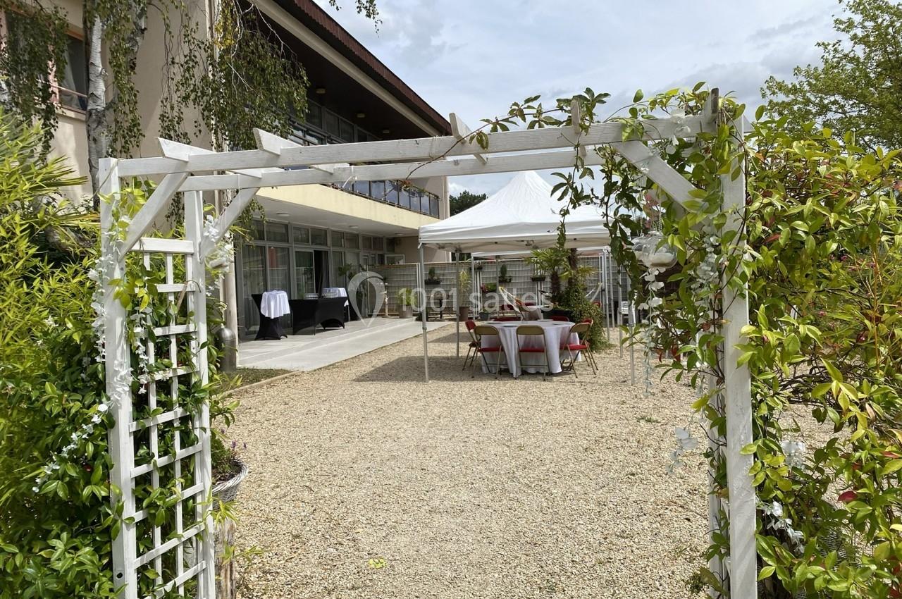 Entrée d'un jardin avec arche blanche, tables dressées sous une tente et bâtiment en arrière-plan.