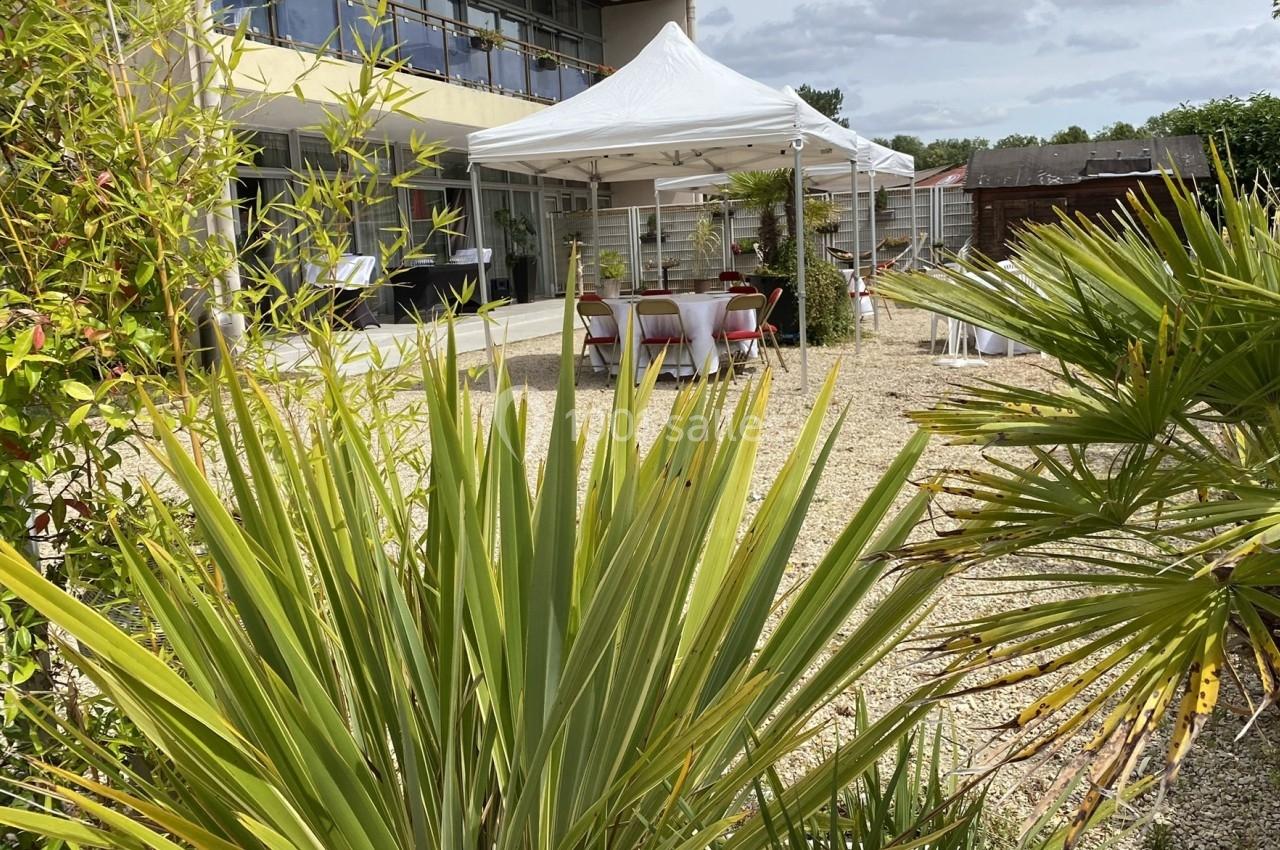 Vue d'un espace extérieur avec des plantes, une terrasse, des tables sous des tonnelles blanches et un bâtiment moderne.