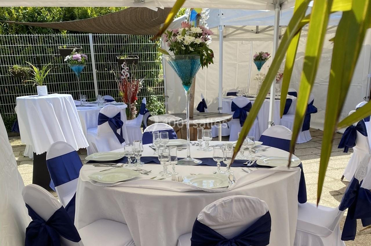 Table dressée pour un événement en extérieur, avec nappes blanches, chaises décorées et compositions florales.