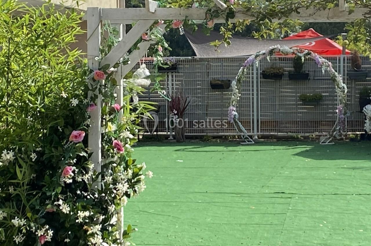 Structure en bois décorée de fleurs roses et blanches, avec une arche florale en arrière-plan sur un sol vert.
