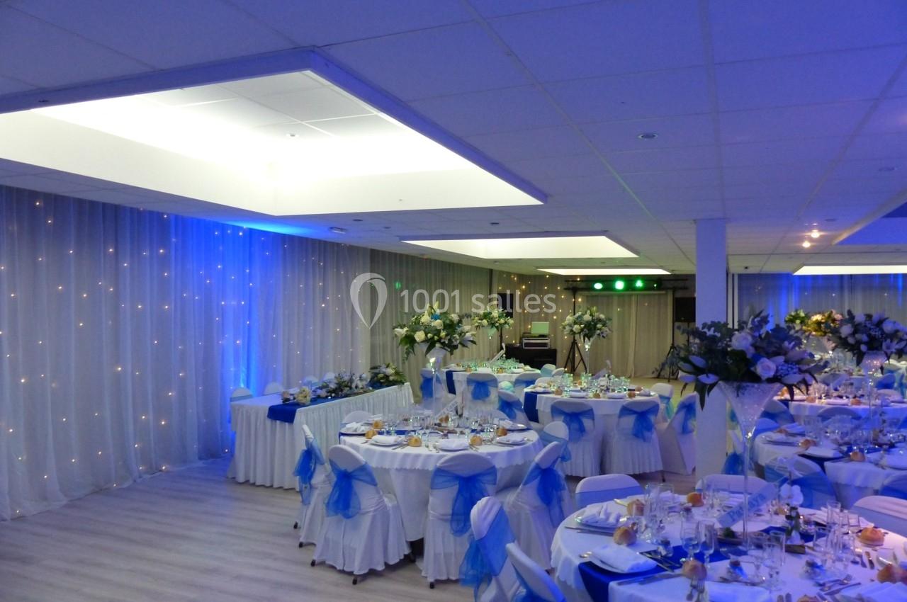 Salle de réception décorée avec des tables rondes, nappes blanches, chaises ornées de nœuds bleus et éclairage tamisé.