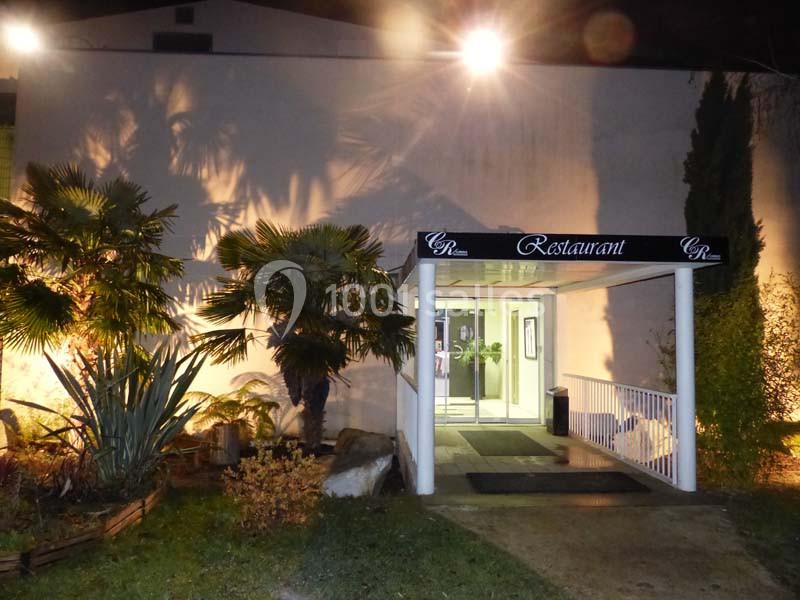 Entrée d'un restaurant éclairée la nuit, entourée de végétation et d'arbustes, avec un auvent noir.