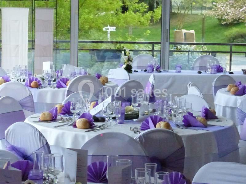 Salle de réception décorée avec des tables rondes, nappes blanches et accents violets, vue sur un extérieur verdoyant.