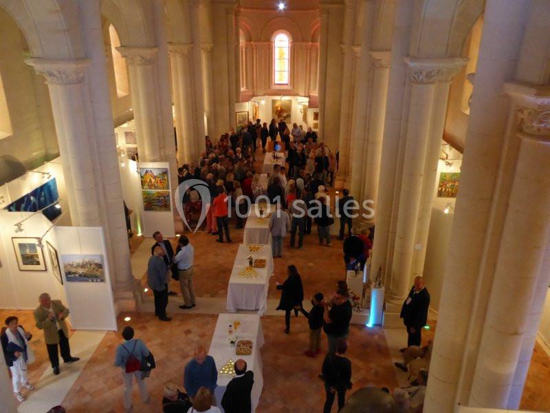 Vue d'une exposition d'art dans une église, avec des visiteurs observant des œuvres et des tables disposées au centre.