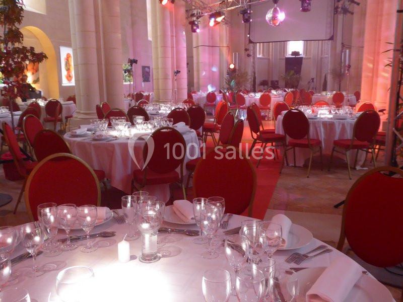 Salle de réception décorée avec des tables rondes dressées, chaises rouges et éclairage tamisé.