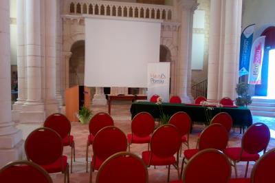 Salle avec des chaises rouges alignées, une table avec des micros, un écran de projection et des bannières promotionnelles.