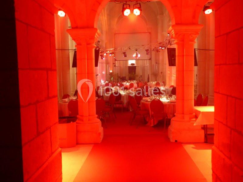 Salle de réception éclairée en rouge, avec des tables dressées et des chaises rouges sous des arches en pierre.
