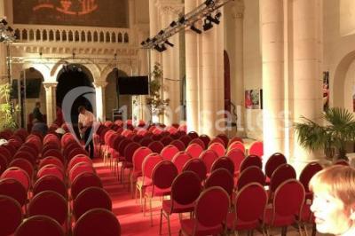 Salle avec des rangées de chaises rouges alignées sur un tapis rouge, entourée de colonnes et éclairée par des projecteurs.