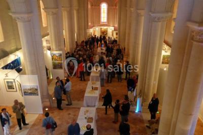 Salle avec des rangées de chaises rouges alignées sur un tapis rouge, entourée de colonnes et éclairée par des projecteurs.