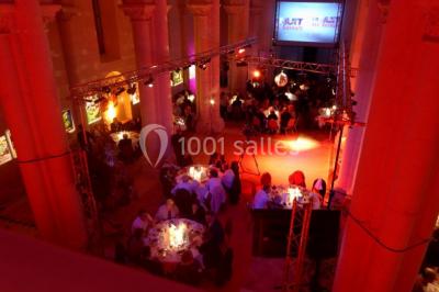 Salle avec des rangées de chaises rouges alignées sur un tapis rouge, entourée de colonnes et éclairée par des projecteurs.
