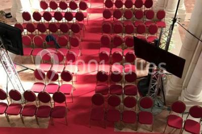 Salle avec des rangées de chaises rouges alignées sur un tapis rouge, entourée de colonnes et éclairée par des projecteurs.