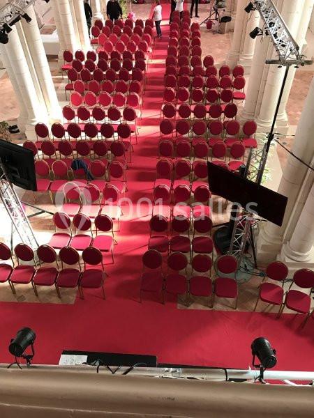 Rangées de chaises rouges alignées sur un tapis rouge dans une salle avec des colonnes et un éclairage de scène.