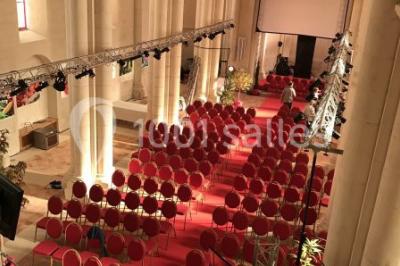 Salle avec des rangées de chaises rouges alignées sur un tapis rouge, entourée de colonnes et éclairée par des projecteurs.