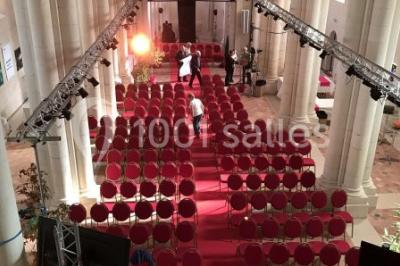 Salle avec des rangées de chaises rouges alignées sur un tapis rouge, entourée de colonnes et éclairée par des projecteurs.