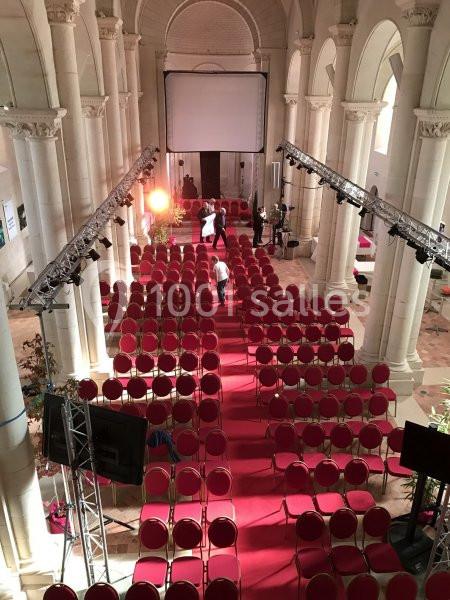 Salle avec des rangées de chaises rouges alignées sur un tapis rouge, éclairée par des projecteurs, dans un bâtiment voûté.