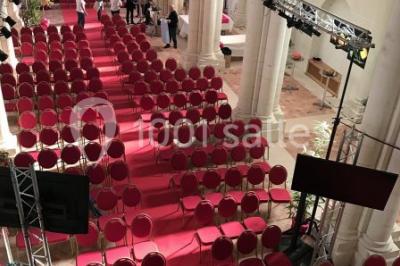 Salle avec des rangées de chaises rouges alignées sur un tapis rouge, entourée de colonnes et éclairée par des projecteurs.