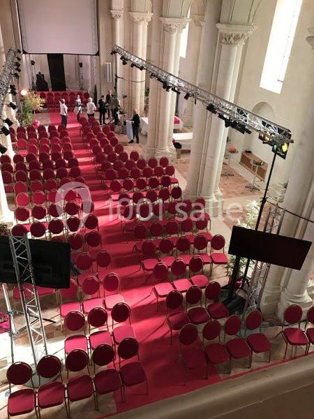 Salle avec des rangées de chaises rouges alignées sur un tapis rouge, entourée de colonnes et éclairée par des projecteurs.