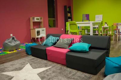 Salle de jeux lumineuse avec une cabane en tissu, un tapis de chiffres, une cuisine pour enfants et des tables colorées.