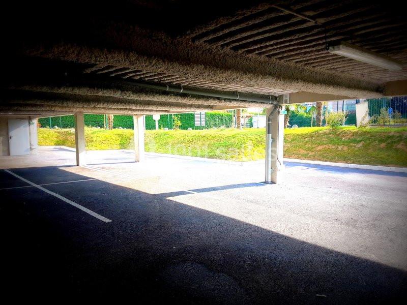 Parking couvert avec sol asphalté, colonnes en béton et lumière naturelle entrant par les côtés ouverts.