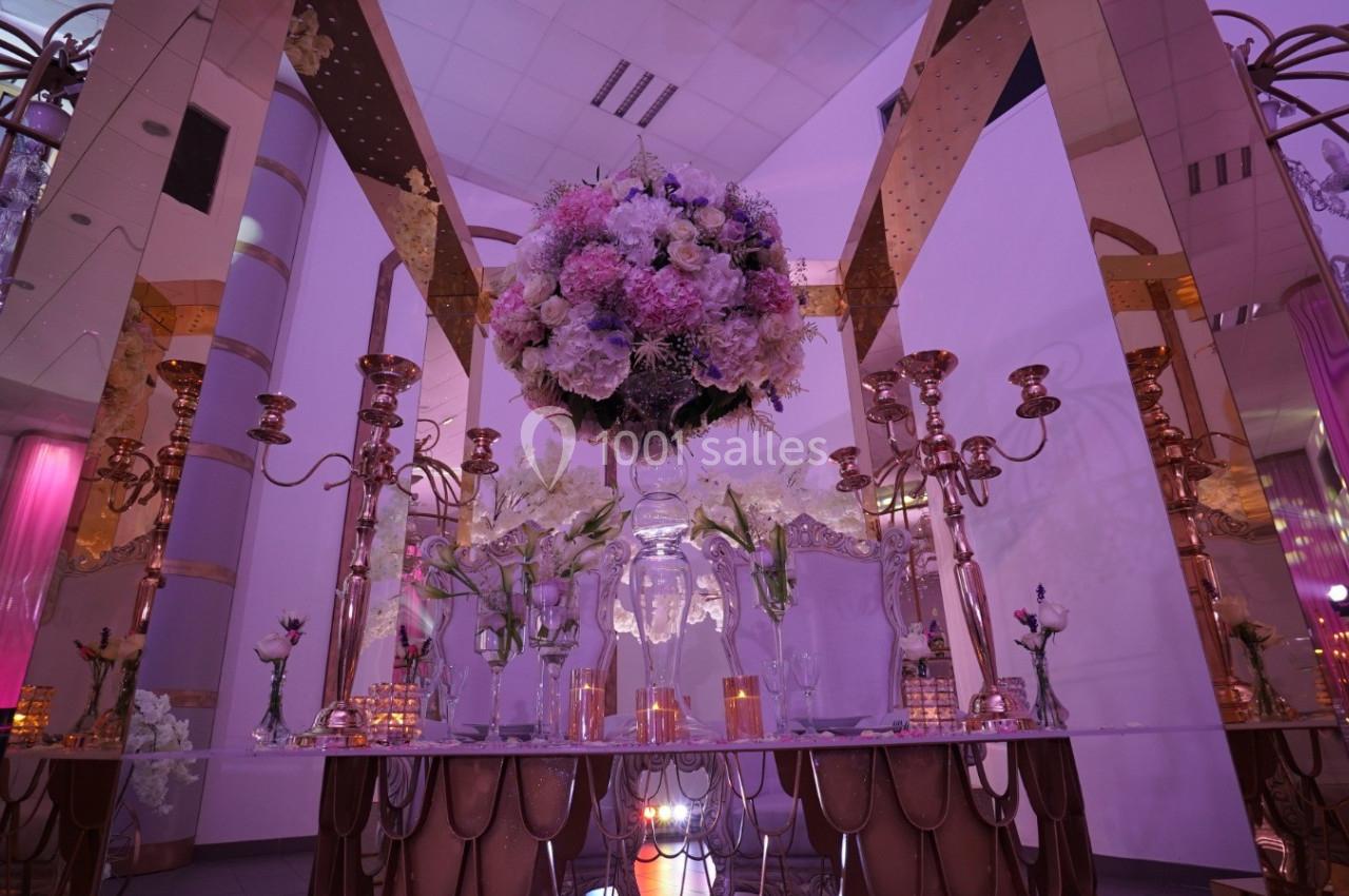 Table décorée avec des chandeliers dorés, des fleurs roses et blanches, dans une salle aux tons violets et dorés.