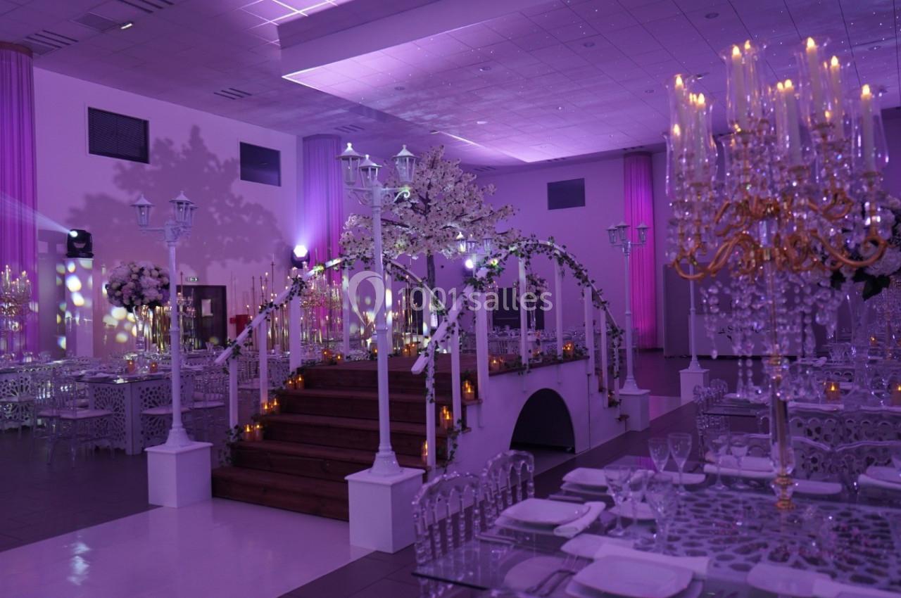 Salle de réception décorée avec un pont illuminé, des chandeliers et des tables dressées sous un éclairage violet.