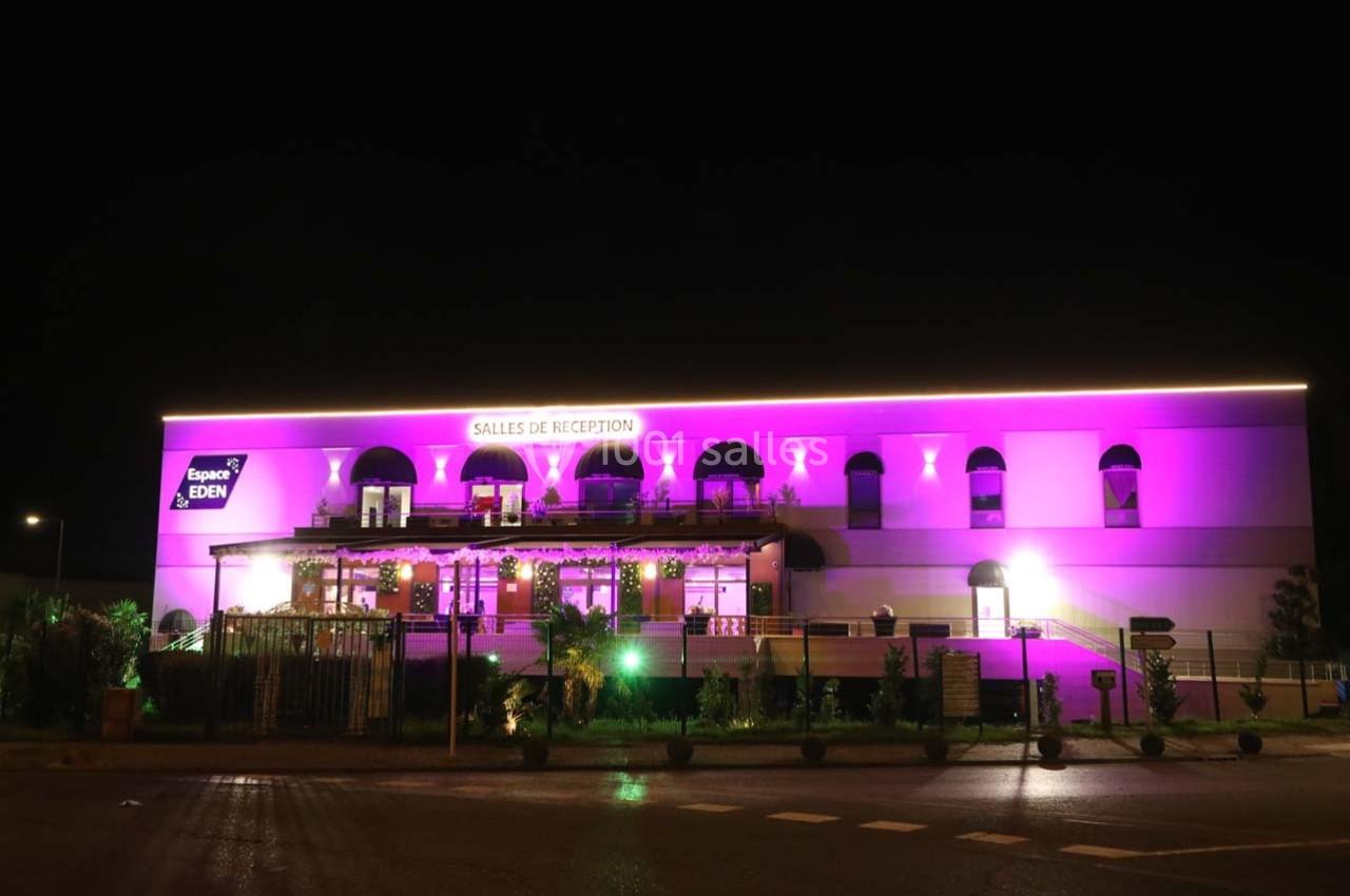 Façade éclairée en violet d'une salle de réception avec terrasse, entourée de végétation, vue de nuit.