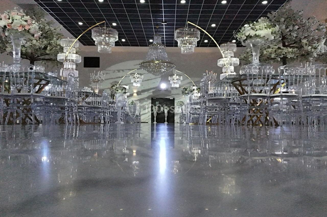 Salle de réception élégante avec sol brillant, tables et chaises transparentes, lustres suspendus et décor floral.