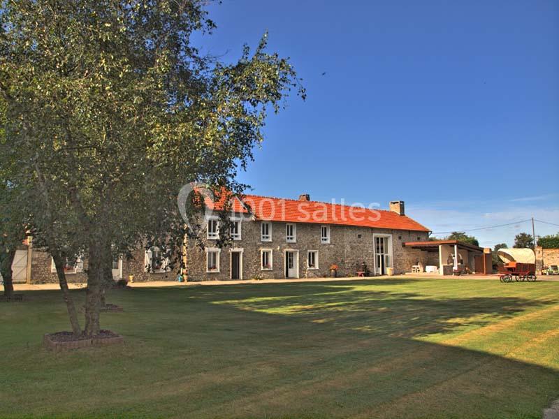 Grande maison en pierre avec toit rouge, entourée d'un vaste jardin arboré sous un ciel bleu clair.