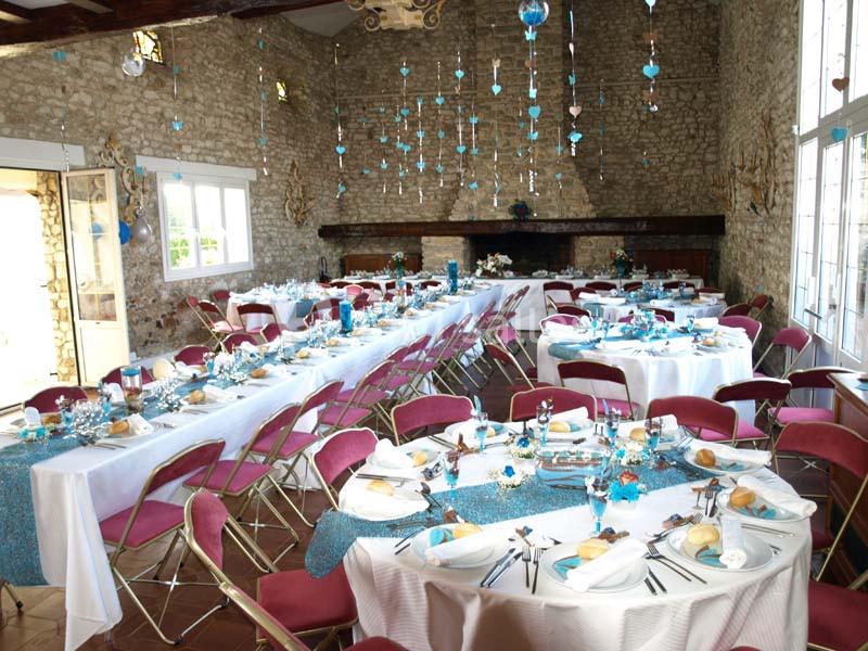Salle de réception décorée avec des tables dressées, nappes blanches et accents bleus, chaises roses pliantes.