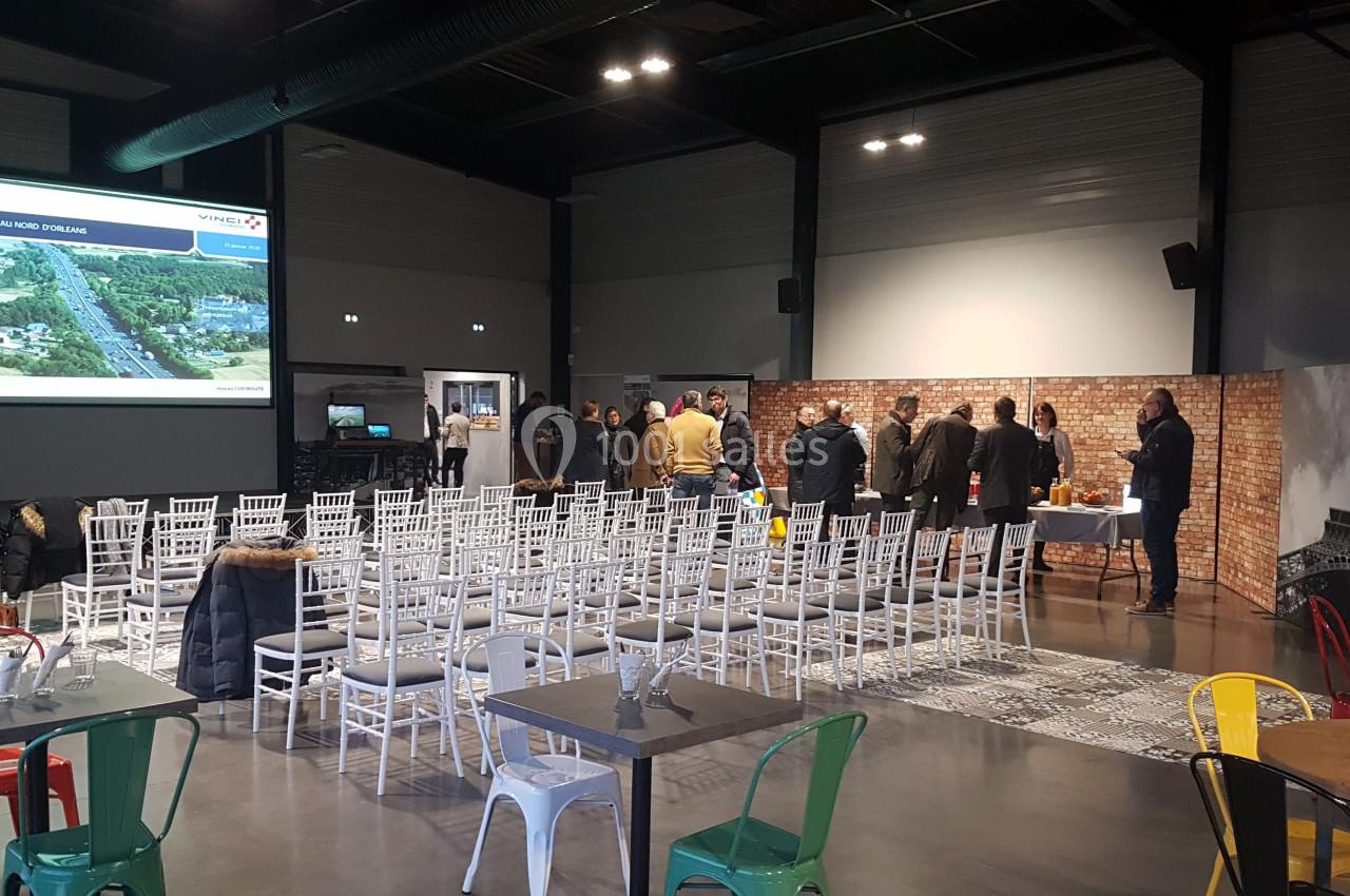 Salle de conférence avec des chaises blanches alignées, un écran de projection et un groupe de personnes discutant au fond.