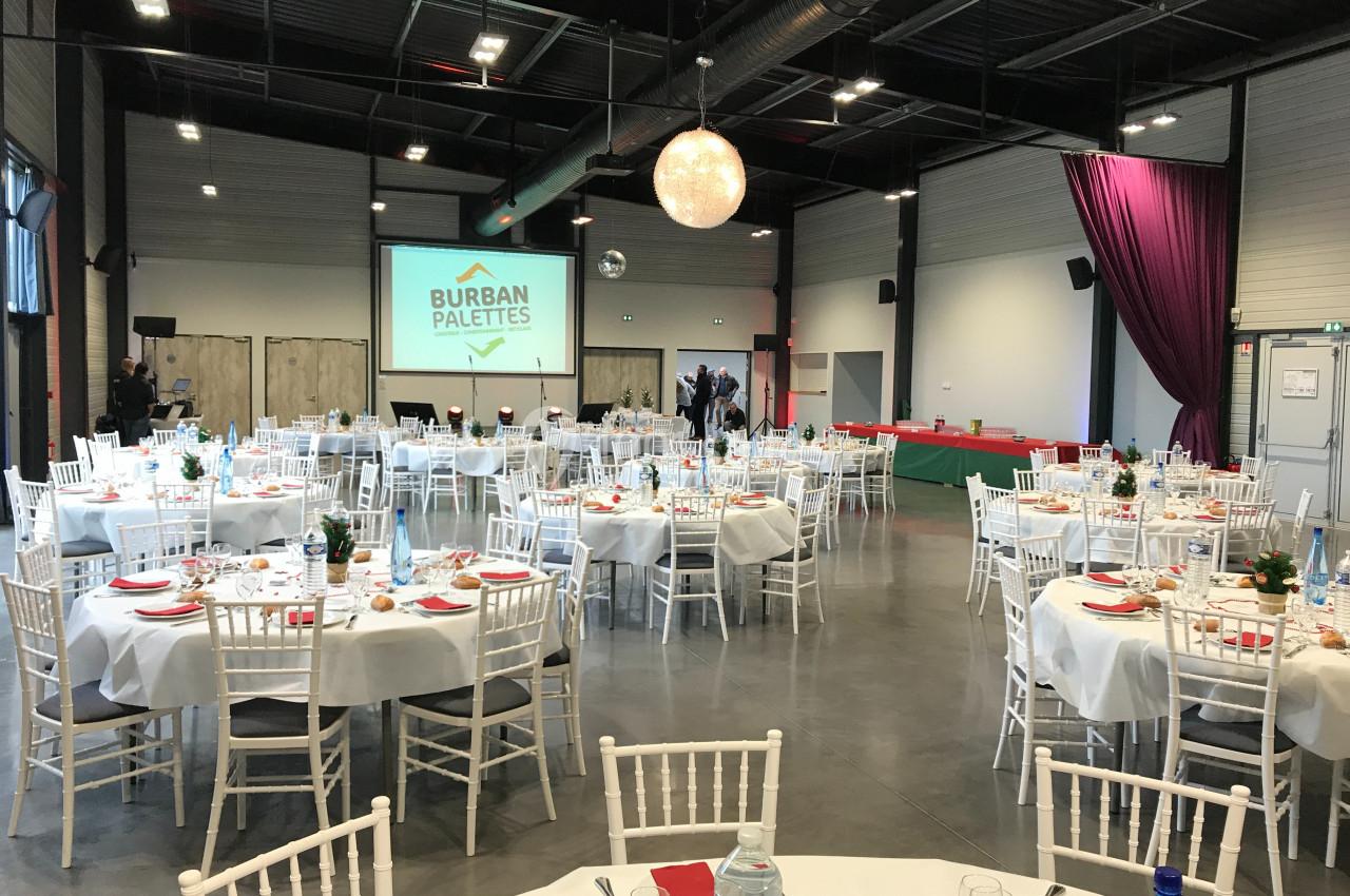 Salle de réception aménagée avec des tables rondes dressées, un écran de projection et une décoration lumineuse.