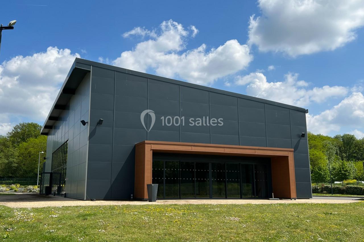 Bâtiment moderne avec façade sombre et entrée en bois, entouré de verdure sous un ciel partiellement nuageux.
