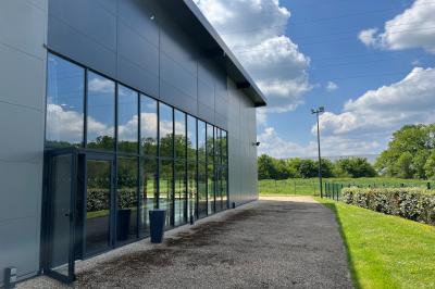 Terrasse avec tables, chaises et parasols, bordée d'une pelouse et d'un bâtiment moderne, sous un ciel ensoleillé.