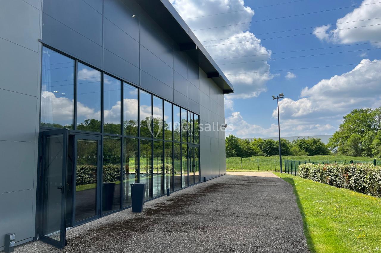 Façade vitrée d’un bâtiment moderne entouré de verdure, sous un ciel partiellement nuageux.
